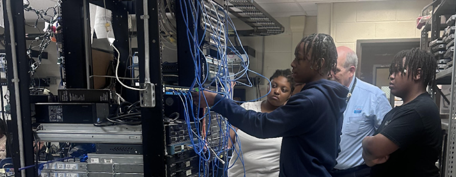 Four people look at a rack of computer equipment.